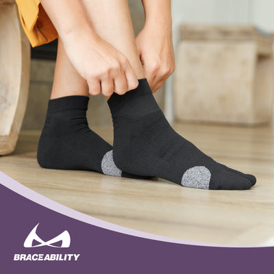 Person wearing black bunion socks with gray cushioning for hallux valgus relief, shown on a wooden floor with BraceAbility logo visible.